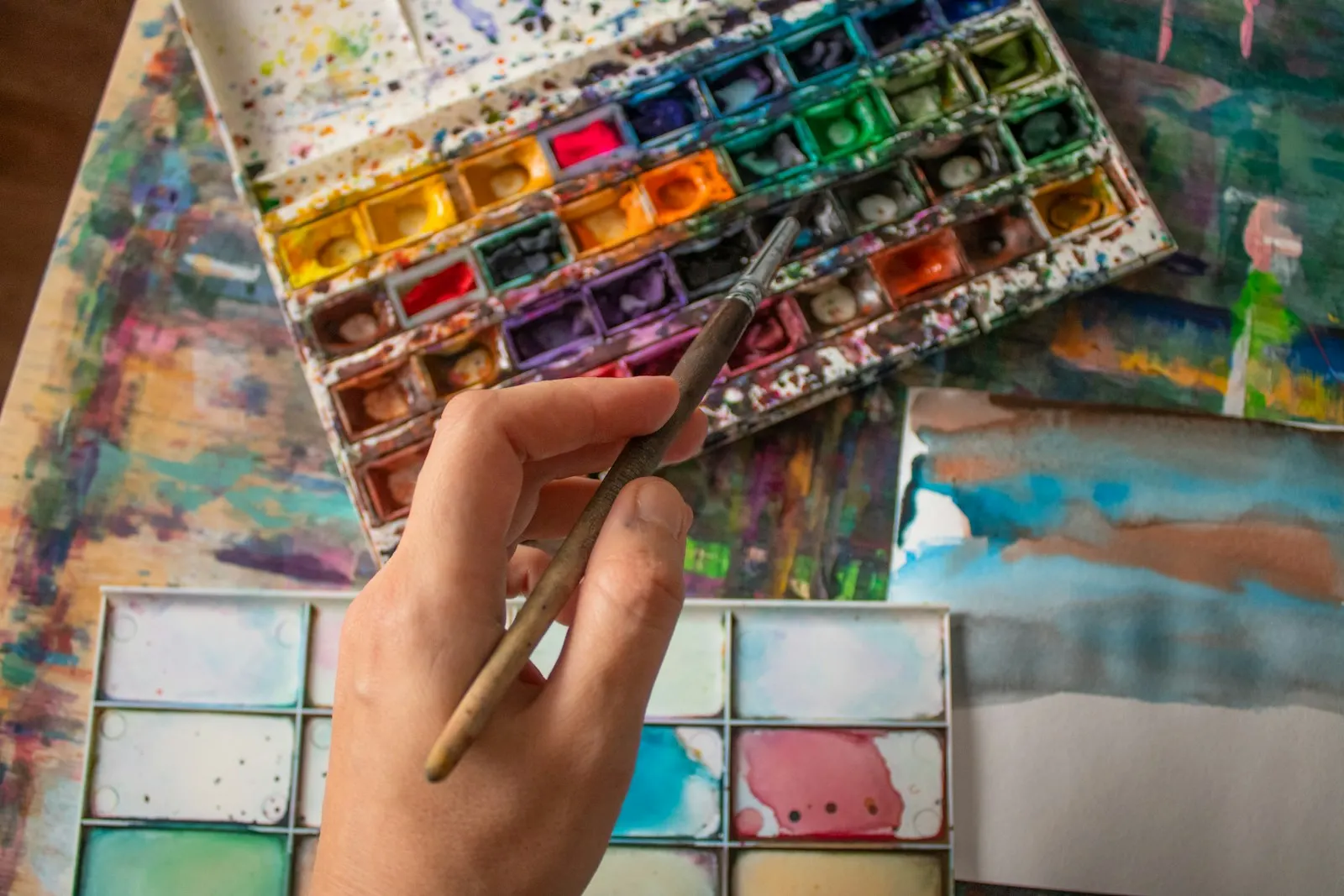 A hand holds a brush over watercolor paints